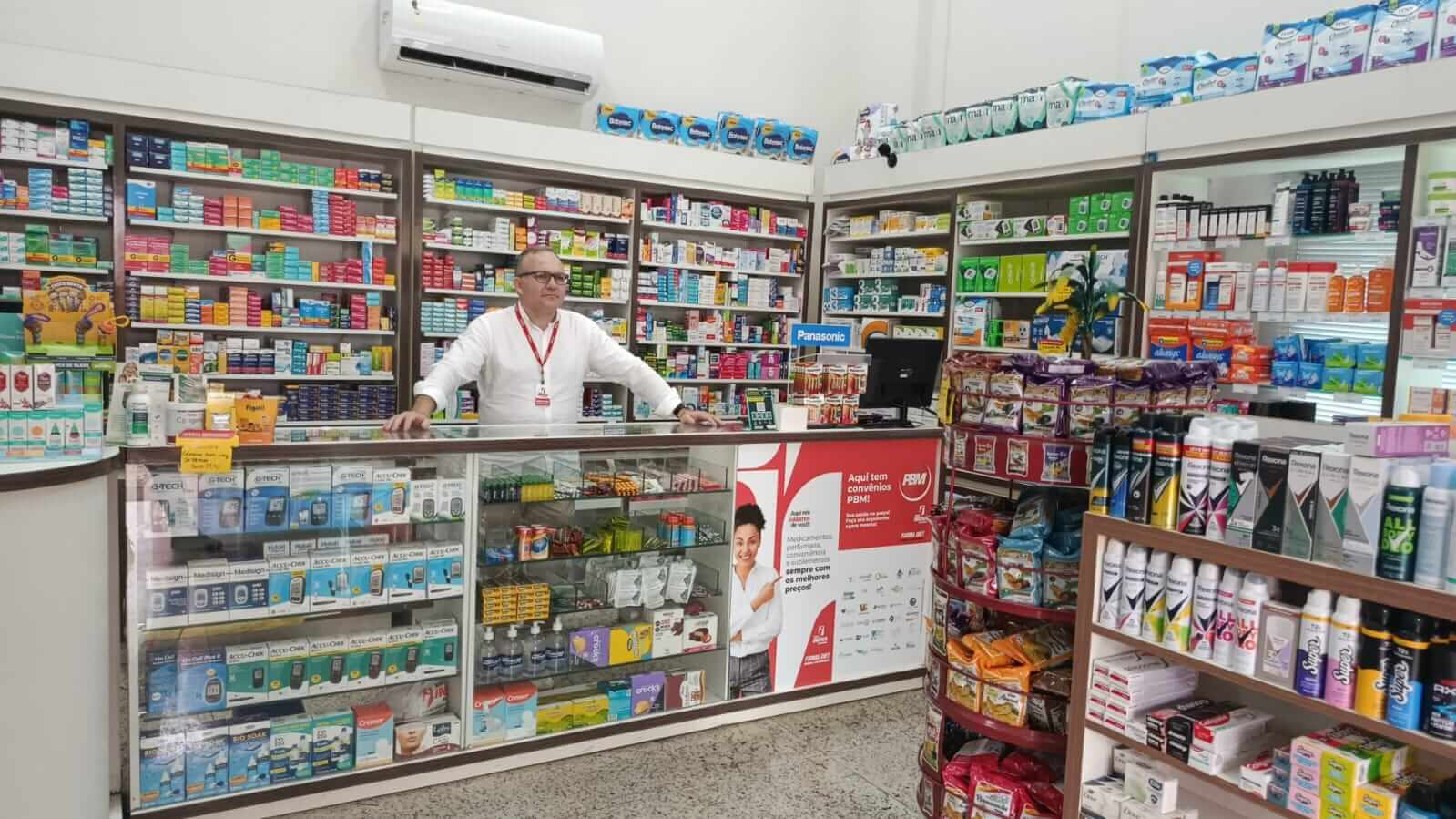 Farmacêutico sorridente atrás do balcão de atendimento da Farma Diet no Centro de Uberlândia, com prateleiras repletas de medicamentos ao fundo